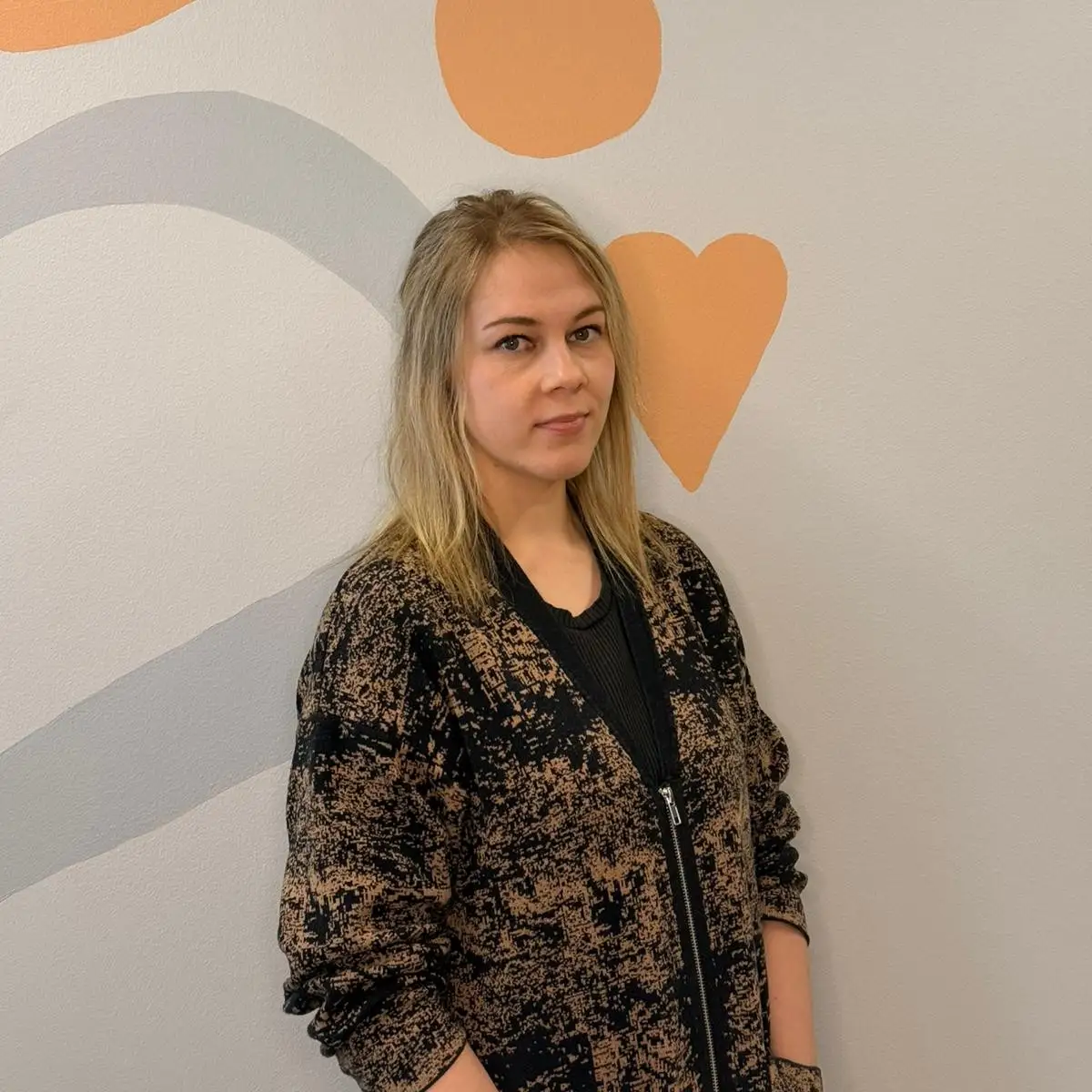 Toimintaterapeutti Ami Alastalo katsoo kameraan kuviollisen seinän edessä.
